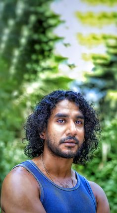 a man with curly hair standing in front of trees and bushes wearing a blue tank top