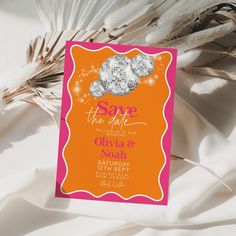 an orange and pink save the date card next to some feathers on a white sheet