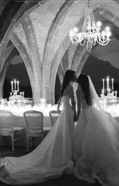 two women in wedding dresses standing next to each other at a table with chandeliers