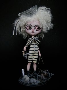 a doll with blonde hair and glasses is standing on a black surface, wearing a striped outfit