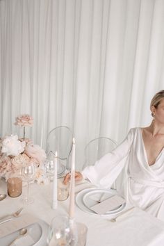 a woman sitting at a dinner table with white linens and flowers in the background