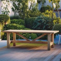 a wooden table sitting on top of a wooden deck next to bushes and trees in the background