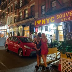 two women are standing in front of a store at night, one is holding her hand on the other's shoulder