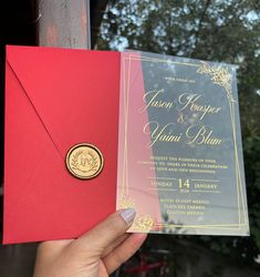 a person holding up a red and gold wedding card with a wax stamp on it