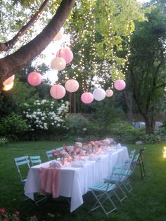 a long table with pink and white paper lanterns hanging from it's ceiling in the backyard