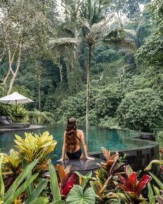 a woman sitting on the edge of a pool surrounded by tropical plants and trees,