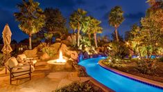 an outdoor fire pit surrounded by palm trees at night with chairs and umbrellas around it