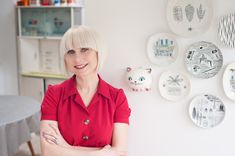 a woman in a red shirt is standing next to plates on the wall and smiling at the camera