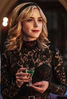 a woman holding a green cocktail in her hand and looking at the camera while wearing a black lace top