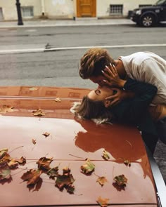 a man and woman kissing on the hood of a car with leaves all over it