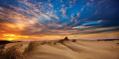 the sun is setting over sand dunes with grass in the foreground and clouds in the background