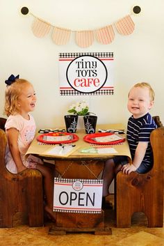 two young children sitting at a table with plates on it and an open sign hanging above them