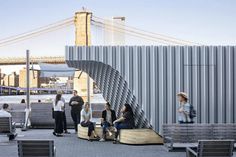 people are sitting on benches near the water and bridge in front of them, while others walk by