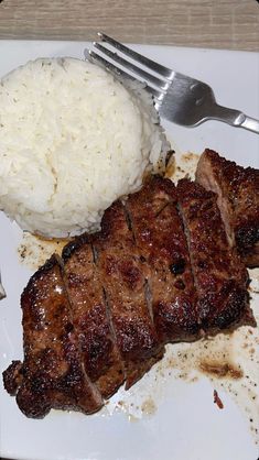 a white plate topped with meat and rice next to a fork on top of a table