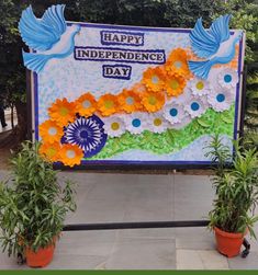 there is a sign that says happy independence day with flowers in the foreground and two potted plants next to it