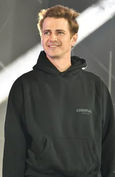 a man in a black hoodie smiles at the camera with his hand on his hip