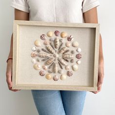 a woman holding a framed artwork with seashells in it's center and shells on the bottom