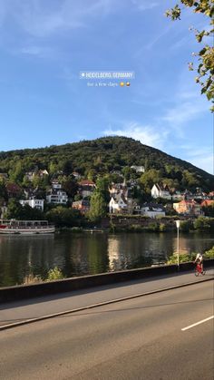 a person riding a bike down a road next to a body of water with houses on the hill in the background