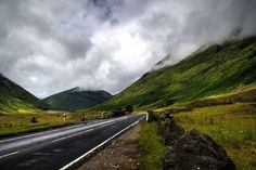 an empty road in the middle of mountains with clouds above it and grass on both sides