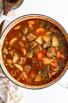 a pot filled with meat and vegetable soup on top of a white tablecloth next to a spoon