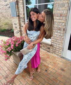 two young women are hugging each other in front of a brick building with pink flowers