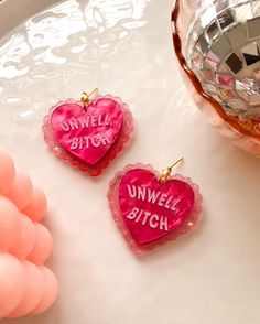 two pink heart shaped earrings sitting on top of a white table next to a vase