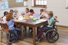 a group of children sitting around a wooden table in front of a woman on a wheel chair