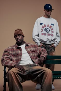 two men are sitting on a bench and one is wearing a baseball cap while the other wears a sweater