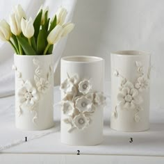 three white vases with flowers in them sitting on a tableclothed surface next to each other