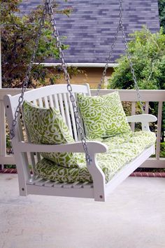 a white porch swing with green and white pillows on the back patio, next to some bushes