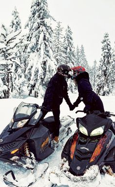 two people on snowmobiles touching hands in the snow with trees in the background