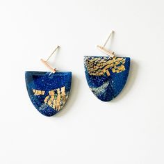 two blue earrings with gold leaf designs hanging from hooks on a white surface, one is shaped like a half - moon