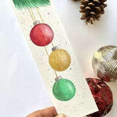 a hand holding up a christmas card with ornaments on it and pine cones in the background