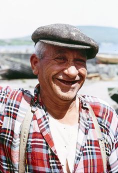 an old man with suspenders and a hat smiling at the camera while standing in front of boats