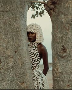 a woman standing between two large trees in front of the ocean, wearing a crocheted headdress