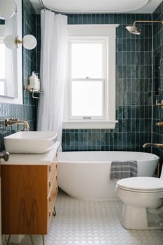 a white toilet sitting next to a bath tub under a window in a green tiled bathroom