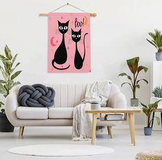 two black cats sitting on top of a white couch in a living room next to potted plants