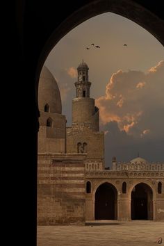 an archway leading to a building with a clock tower in the background and birds flying overhead