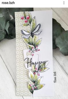 a card with the word happy written on it and flowers in front of it, next to some green leaves