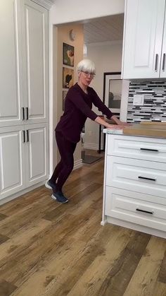 an older woman standing in the middle of a kitchen with white cabinets and wood floors