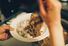 a person holding a spoon over a bowl filled with rice and beef stew on top of a stove