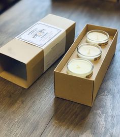 three candles in a cardboard box sitting on a wooden floor next to an open package