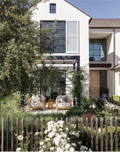 a white house with lots of windows and flowers in the foreground, next to a wooden fence