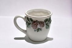 a white coffee cup with pine cones and leaves painted on the side, sitting on a white surface