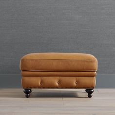 a tan leather footstool sitting on top of a hard wood floor next to a gray wall