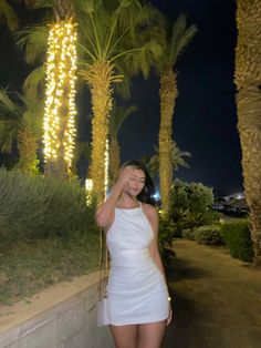 a woman in a white dress is talking on her cell phone while standing next to palm trees