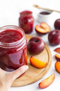 a person holding a jar of plum jam with sliced peaches around it on a cutting board