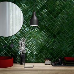 a green tiled wall with a round mirror and vase on the table next to it