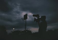 a man holding a camera in front of a basketball hoop at night with the moon behind him