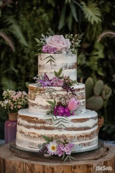 a three tiered cake with flowers and greenery on top sits on a tree stump